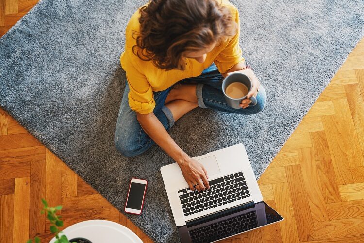 young girl woman in a yellow shirt works on a laptop with a mug of coffee at home in the living room on the carpet, remote work and education. Top view